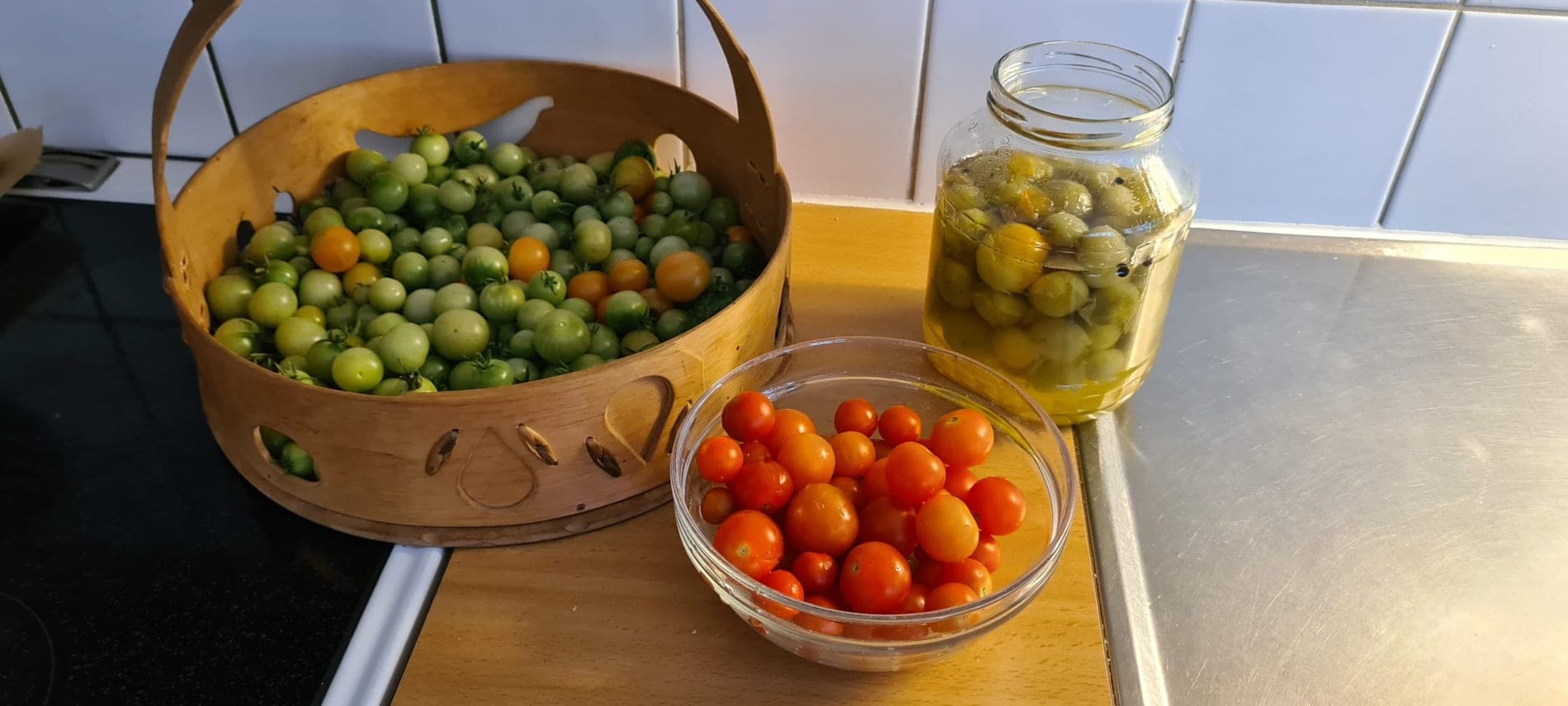 Nyskördade tomater i en näverkorg och en glasburk med inlagda gröna tomater på köksbänken.