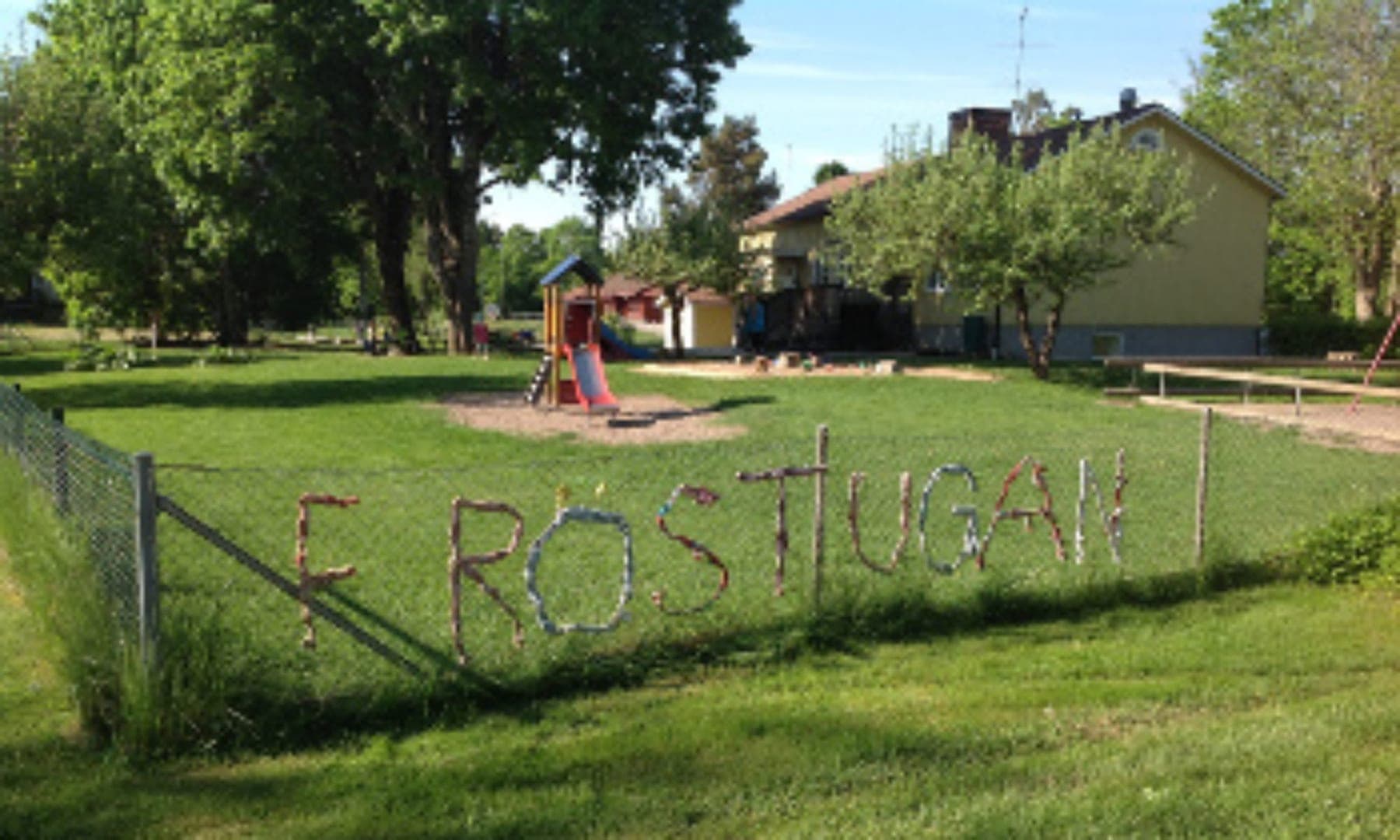 Gården vid huvudbyggnaden en sommardag, med lekställning, sandlåda och stora lövträd.