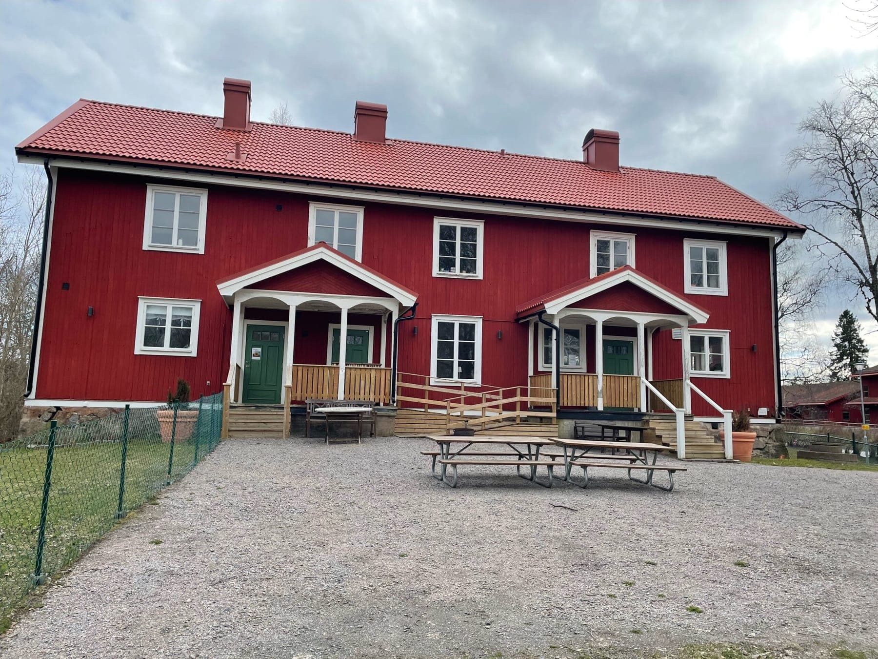 Den gamla kyrkskolan i Riala – en falurödmålad tvåvåningsbyggnad med vita knutar, gröna dörrar och picknickbord på grusplanen.